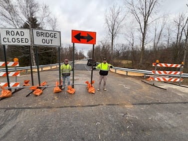 Two workers holding closure signs on the 32nd Avenue bridge over the north branch of Crockery Creek in Ottawa County. 