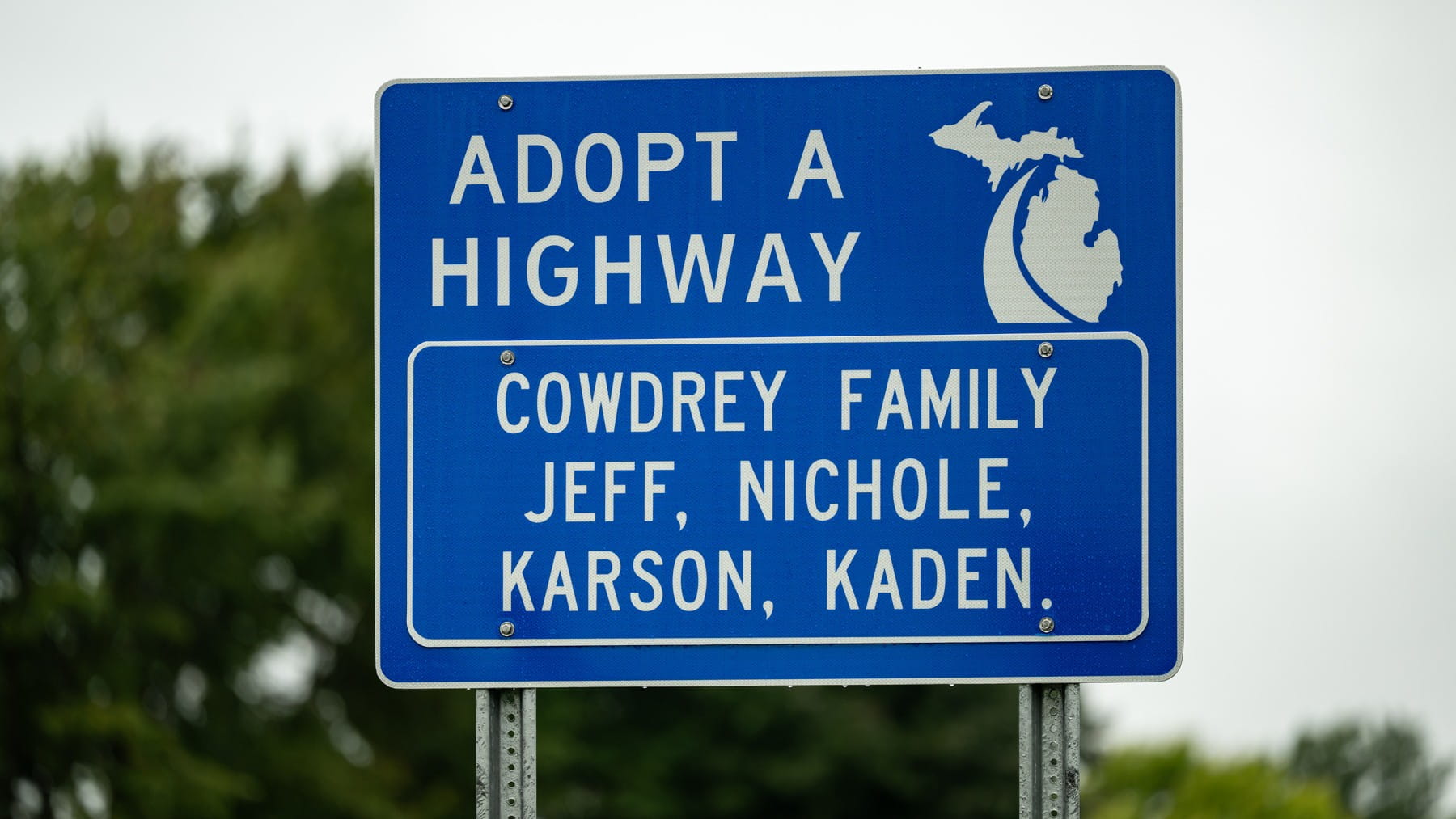 Blue Adopt-A-Highway sign featuring MDOT's swoosh logo and the Cowdrey family names.