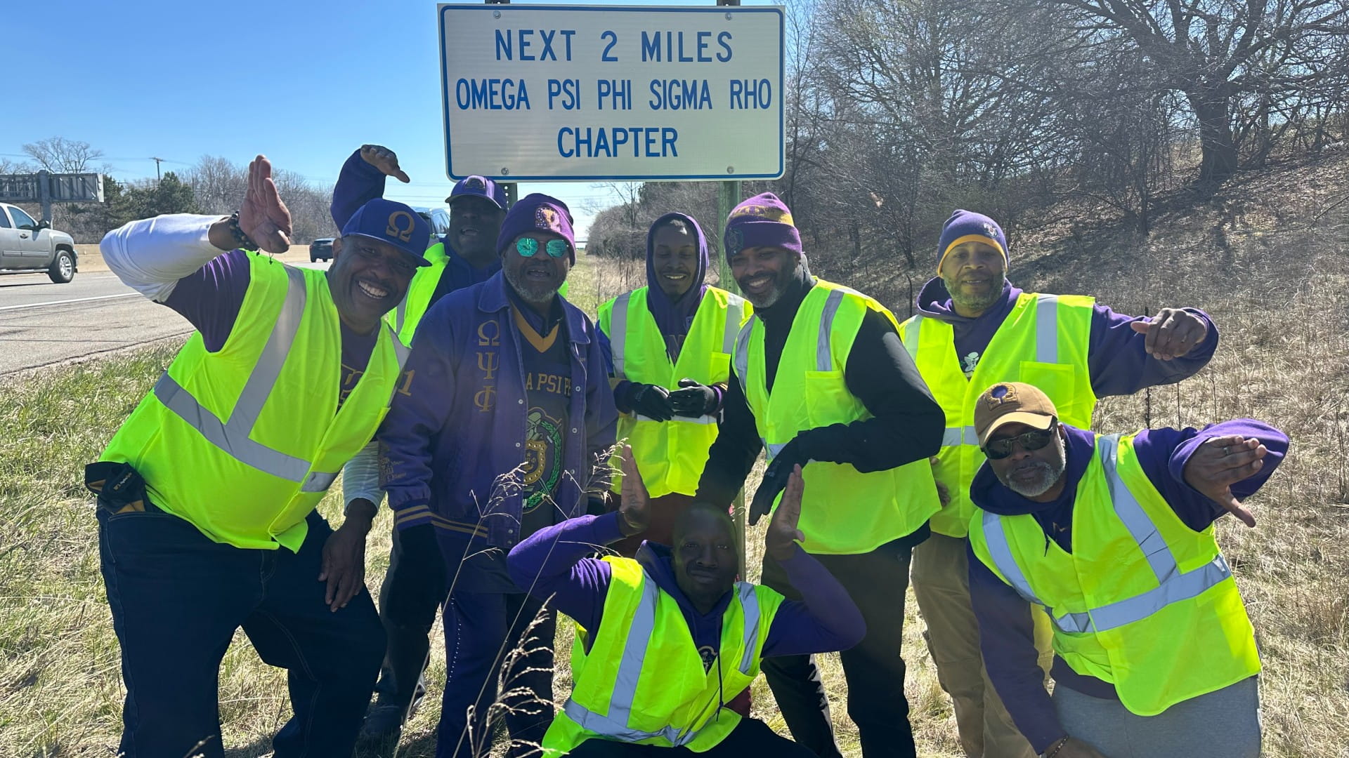 Group of Adopt-A-Highway (AAH) volunteers wearing high‑visibility safety vests gathered beneath an AAH sign for a group photo. 