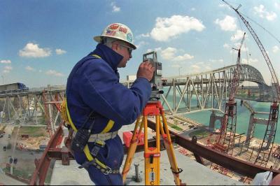 An employee at a bridge construction site by a river using surveying equipment.