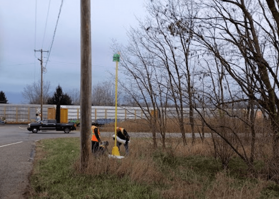 Two workers in reflective vests install a wireless communications unit on the side of a rural road.