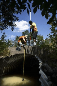 Two people on a bridge installing water-monitoring equipment into the water.