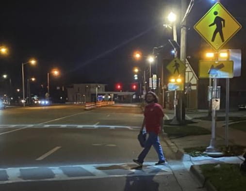 A pedestrian walking in a lighted crosswalk in an urban nighttime setting.