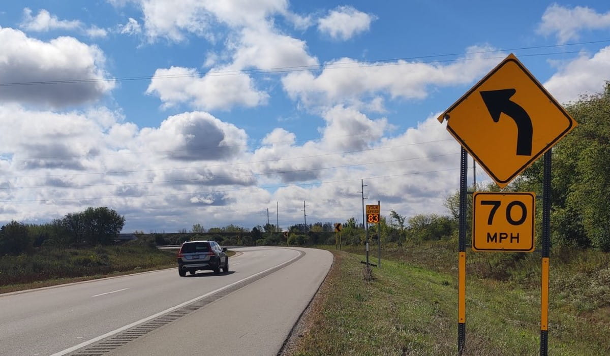 A vehicle passing a sign indicating the speed limit is 70 mph around a curve, while a dynamic speed feedback sign indicates the vehicle’s speed is 83 mph.