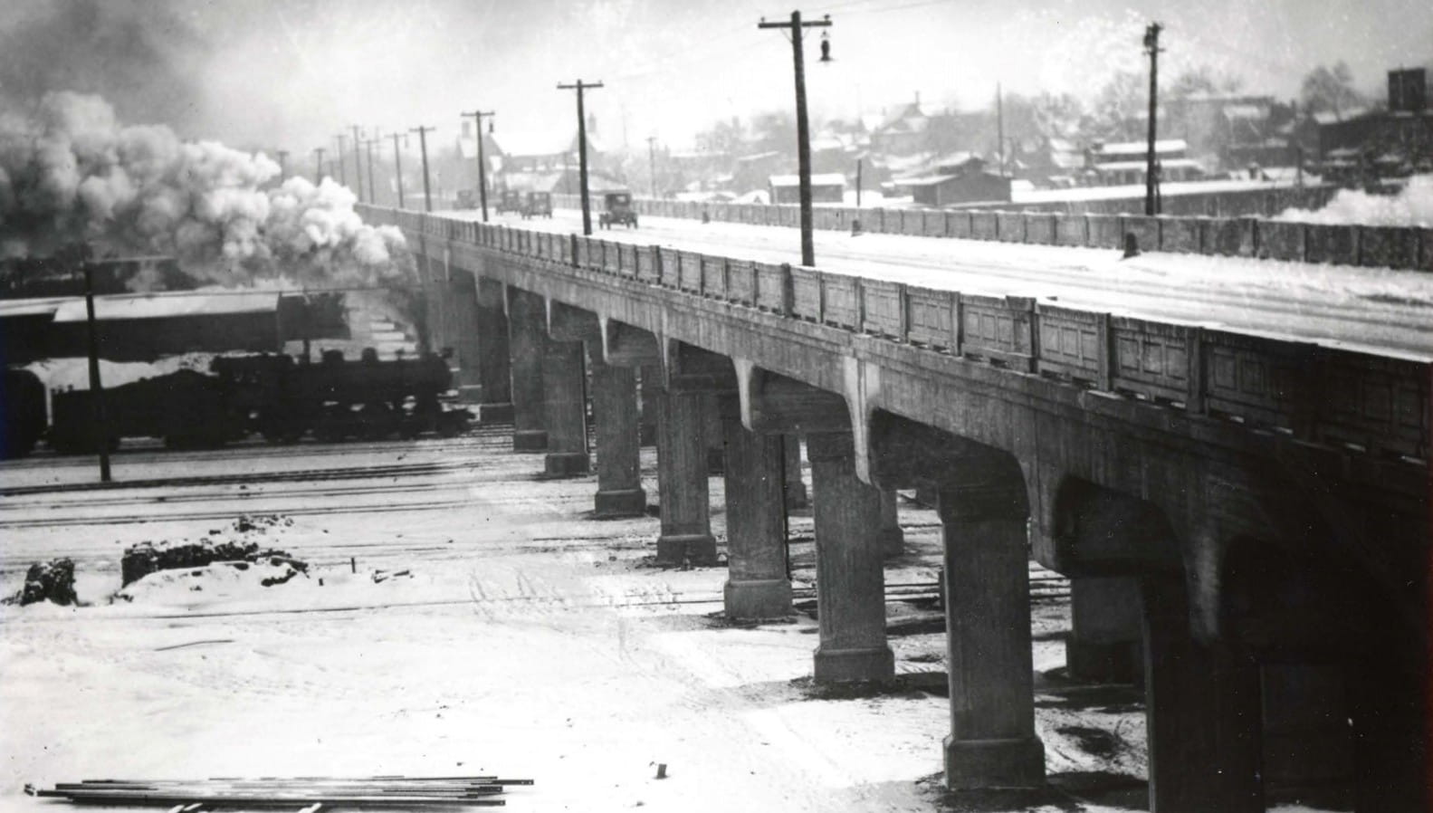 A train travels along multiple railroad tracks toward the Franklin Street viaduct in 1925, as several Model T cars pass overhead. Photo credit: Engineering Department, “Franklin Street Viaduct - Franklin Street Bridge,” City of Grand Rapids Archives and Records Center, accessed Feb. 17, 2026.