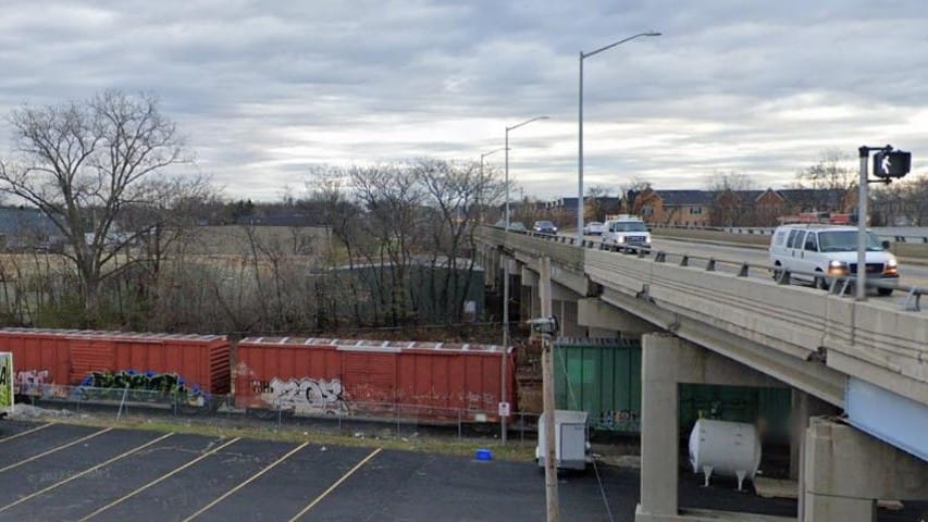 The Franklin Street bridge today, with modern vehicles crossing above active railroad tracks, a passing train below, and a parking lot with semitrailers. Photo credit: Google Maps Street View.
