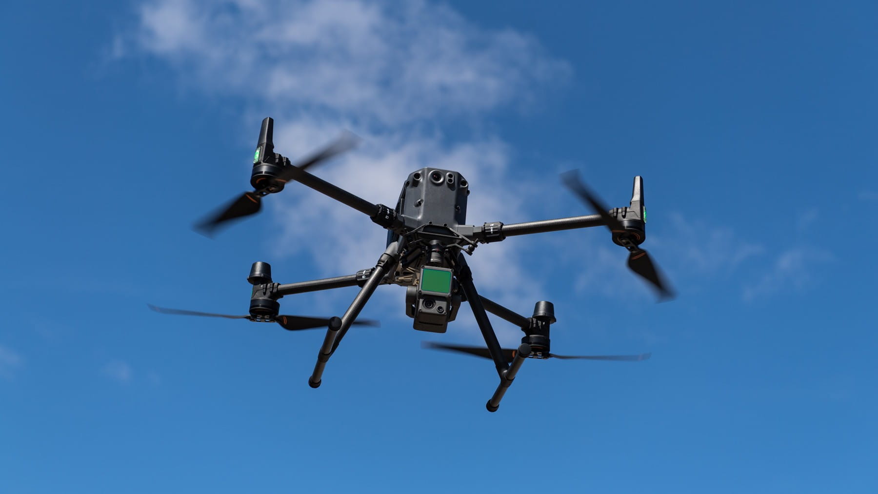 A close up of a drone in flight against a bright blue sky with white wispy clouds.
