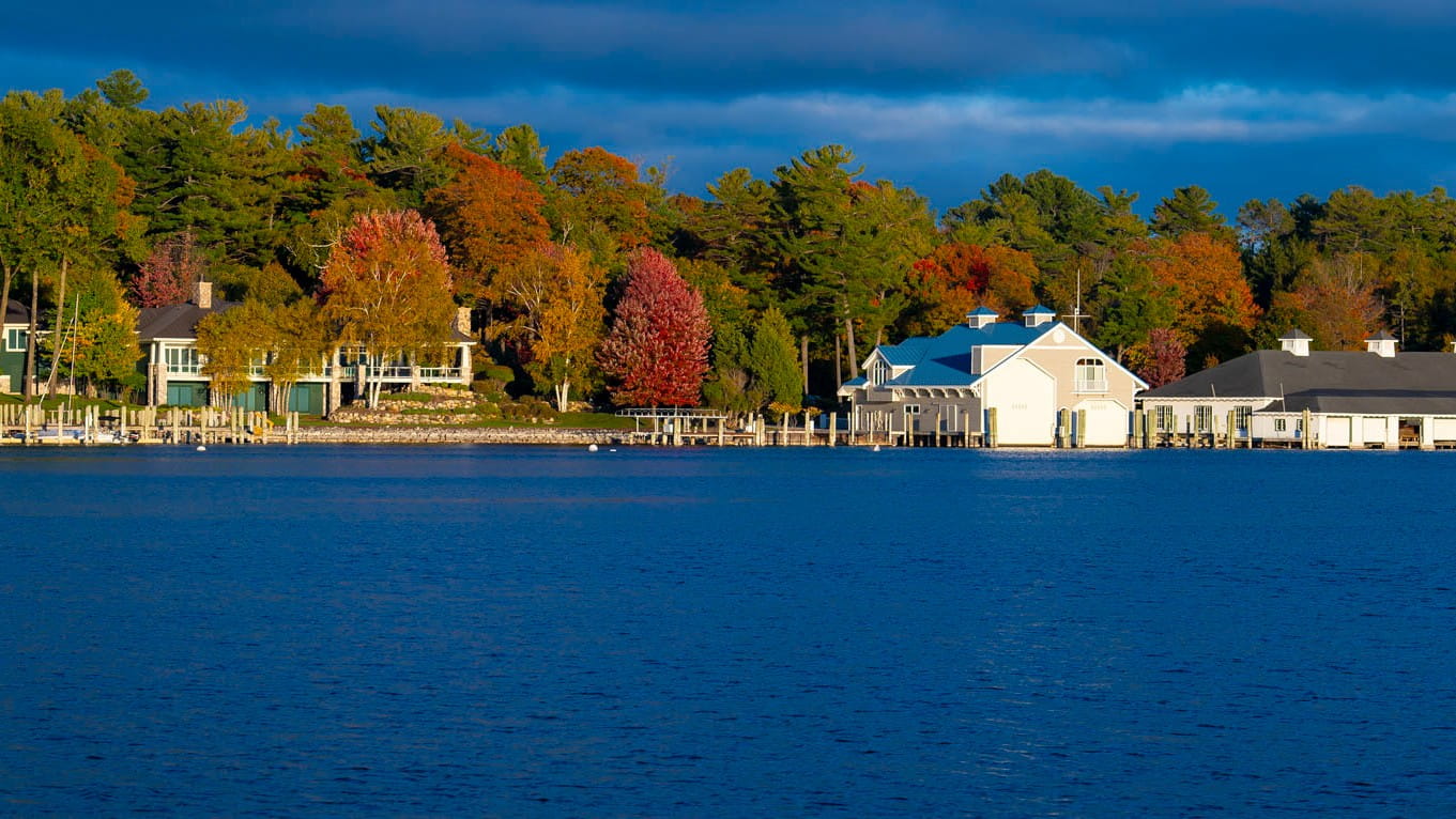 The blue waters of Lake Charlevoix against a dark blue sky with trees in shades of greens, reds and oranges on a early autumn morning.