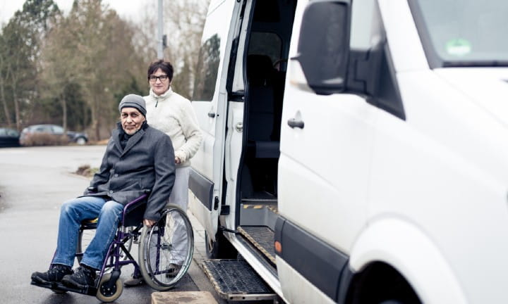 A woman with a gentleman sitting in a wheelchair next to a small bus.