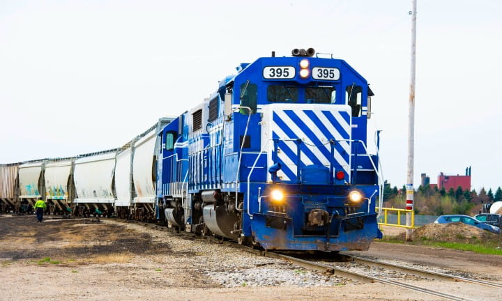 A blue train engine with its lights on pulling rail cars.