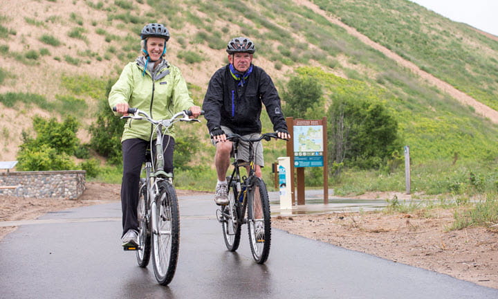 Two bicycle riders wearing helmets while riding along a trail near the Sleeping Bear Heritage Trail.