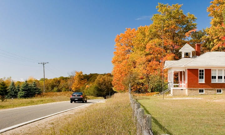 A black vehicle travels along M-119 Tunnel Of Trees on a sunny, autumn day passing the 5 Mile Creek Road School House in Emmet County.