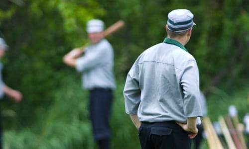 Green trees behind him, player in green-and-black old-time uniform at homeplate ready to swing bat