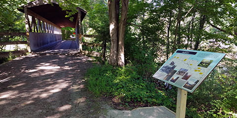 A shady trail features a sign on a post and a covered bridge.