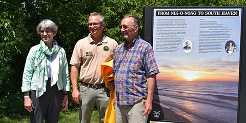 A woman and two men smile while standing in front of a large sign.