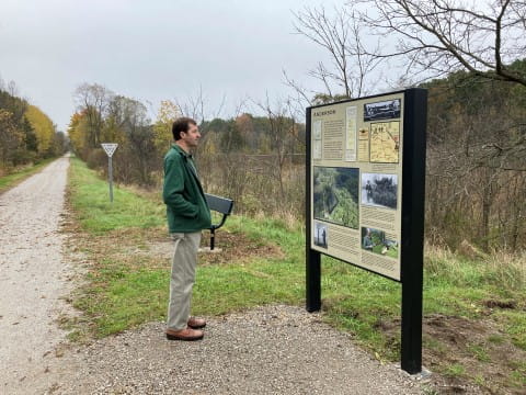 A man in a green jacket stands reading an interpretive sign