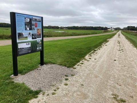 Interpretive sign in a black metal frame, located along a trail which runs off into the distance. A flat and green farm field is in the background