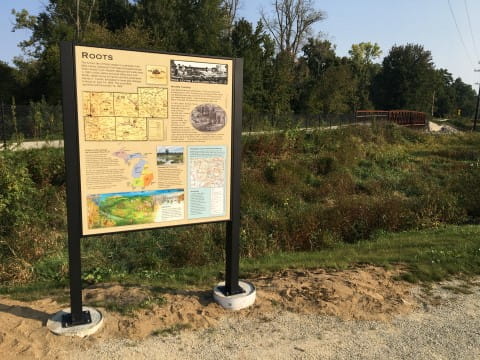 Interpretive sign in a black metal frame, with trees and a steel bridge for the trail in background