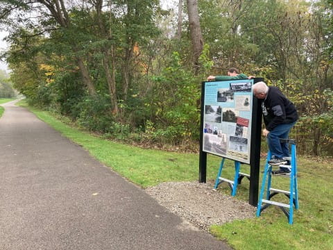 Two men stand on ladders to install an interpretive sign into a metal frame