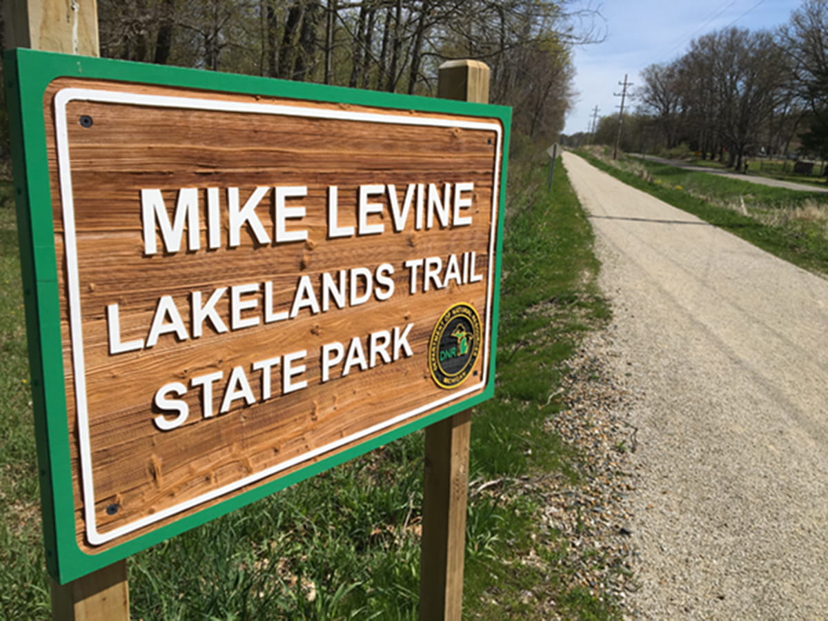A wood sign announces the Mike Levine Lakelands Trail, a trail bordered by trees and a road