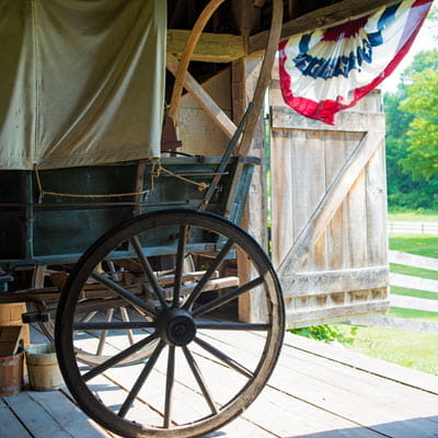 A covered wagon sits inside the open doors of a large brown wooden barn. 