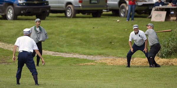 A uniformed man with a baseball bat waits for a flying baseball to reach him. The other men stand around, watching. 