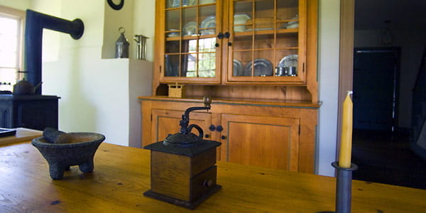 A coffee grinder, mortar and pestle sit on a wooden dining table. A wooden china cabinet is in the background. 