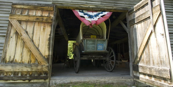 A covered wagon sits inside the open doors of a large brown wooden barn. 