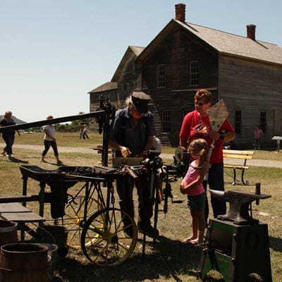 A blacksmith talks to a young girl and a woman on the grass outside a old wooden building.