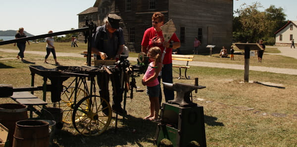 A blacksmith talks to a young girl and a woman on the grass outside a old wooden building.