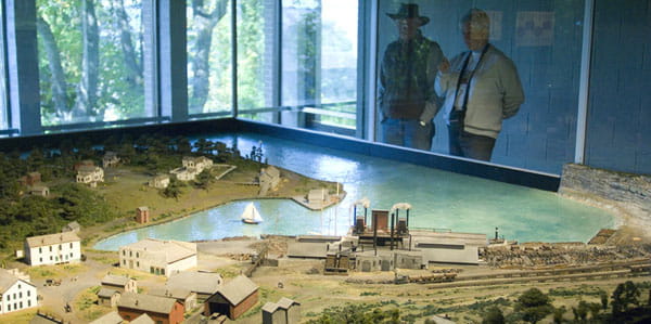 Two people look through a large glass pane at a scale model of a historic industrial town.