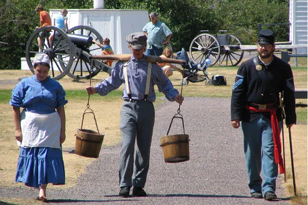 A boy carrying two wooden buckets with a yoke walks a gravel path with a man and a woman. All are in historic costume.