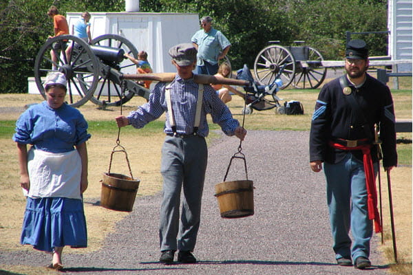 A boy carrying two wooden buckets with a yoke walks a gravel path with a man and a woman. All are in historic costume.