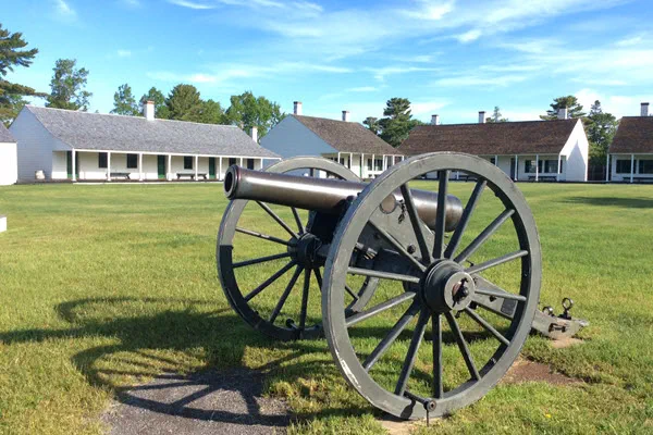 A large, wheeled cannon sits on a large grassy area. White one-story wooden buildings are in the far background.
