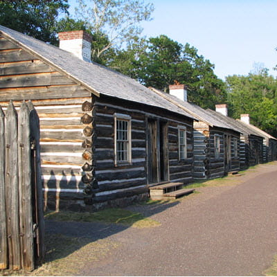 A row of four one-story log cabins line a dirt road. 