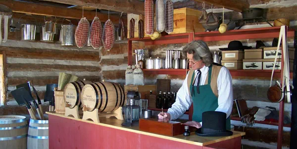A man in an apron stands behind the counter in a log general store. 