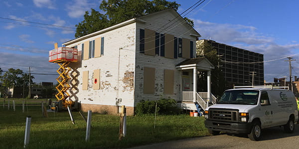 A construction lift sits along the side of a two-story white clapboard home with peeling paint.