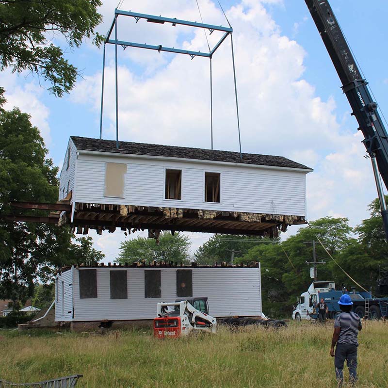 A crane lifts the second story of a white clapboard home off the first story.