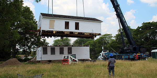 A large crane lists the second story of a two-story clapboard home off the first story. 