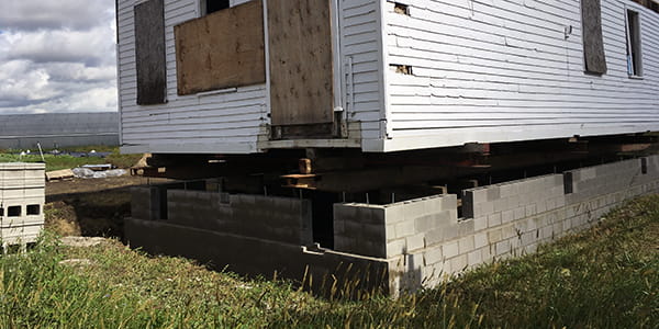The first floor of a white clapboard home is slowly being lifted onto a new cinder block foundation