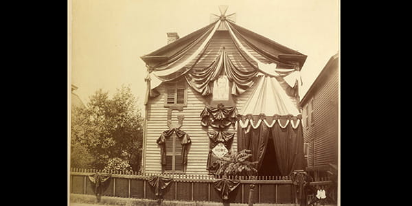 A sepia toned historic image of a two-story clapboard home, draped in swaths of dark fabric..