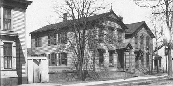 A two-story clapboard home with light walls and dark trim sits behind a picket fence, circa 1906.