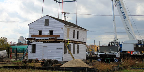 A crane gently places the second story of a two-story white clapboard house onto the first story. 