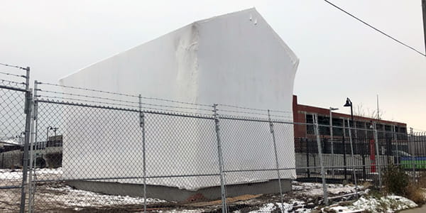 A two story home is shrink-wrapped from top to bottom in opaque gray-white plastic. 