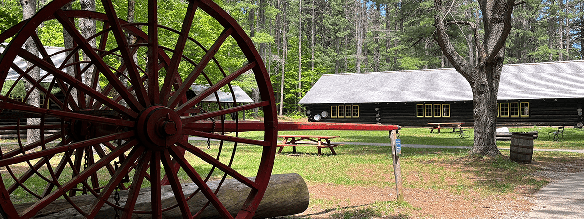 A big red wheel with a log cabin type building in the background, surrounded by trees.