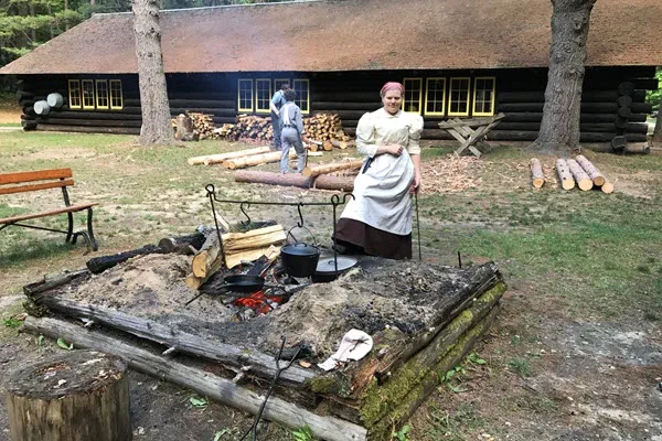 A women in historical clothing stands in front of a large outdoor fire. A pot is suspended and a skillet sits on a grate above the fire.