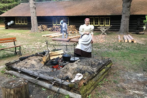 A women in historical clothing stands in front of a large outdoor fire. A pot is suspended and a skillet sits on a grate above the fire.
