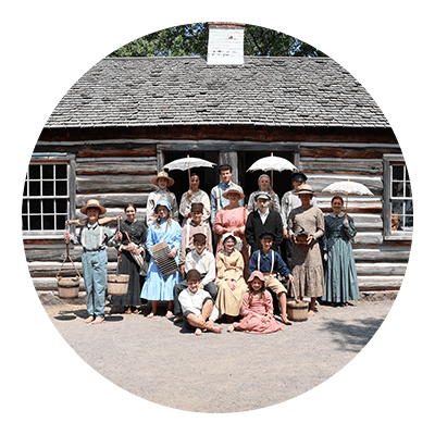 A group of students dressed in reproductions of 1870 style clothing in front of a log cabin.