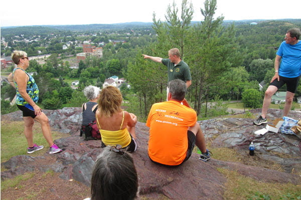 Several people sit on rocks on top of a hill. A man in a green shirt points to the town below. 