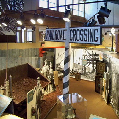 A large black and white railroad crossing pole in the foreground; a mining cart and cut-outs of miners in the background. 
